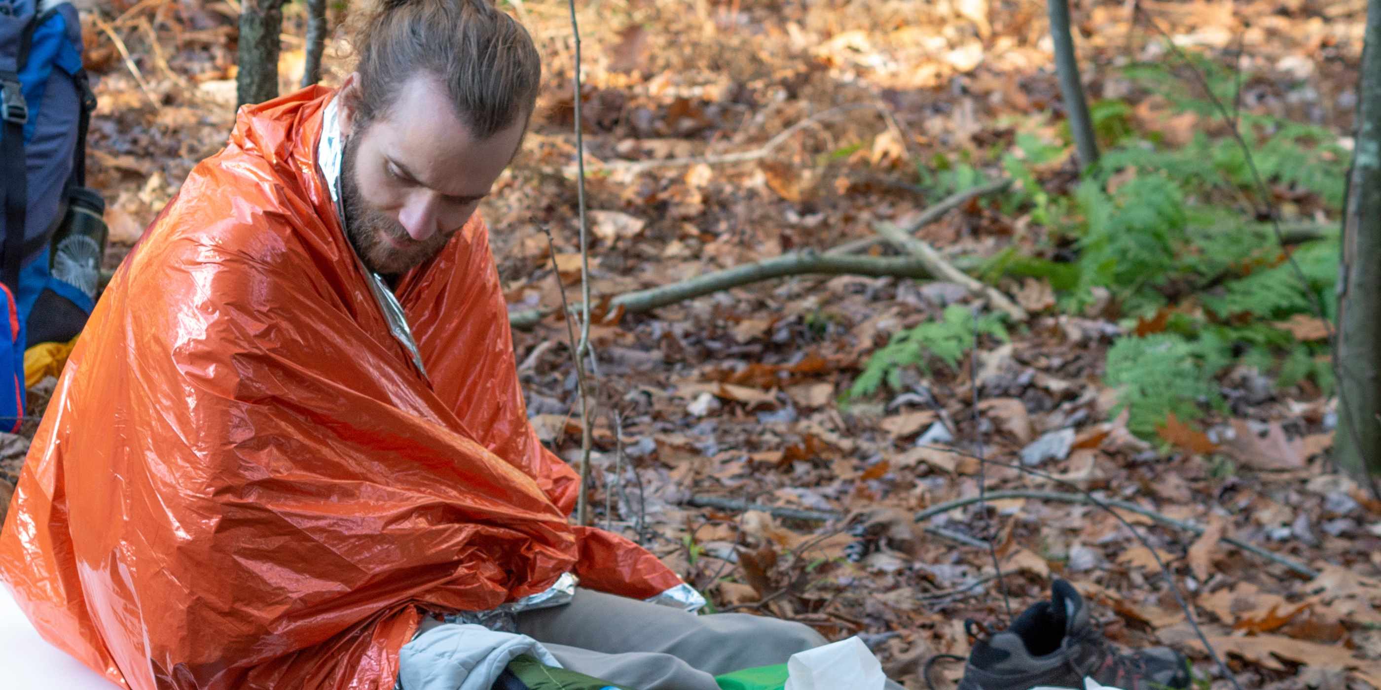 Man Wrapped in Orange SOL Emergency Bivvy Sitting on Leafy Ground