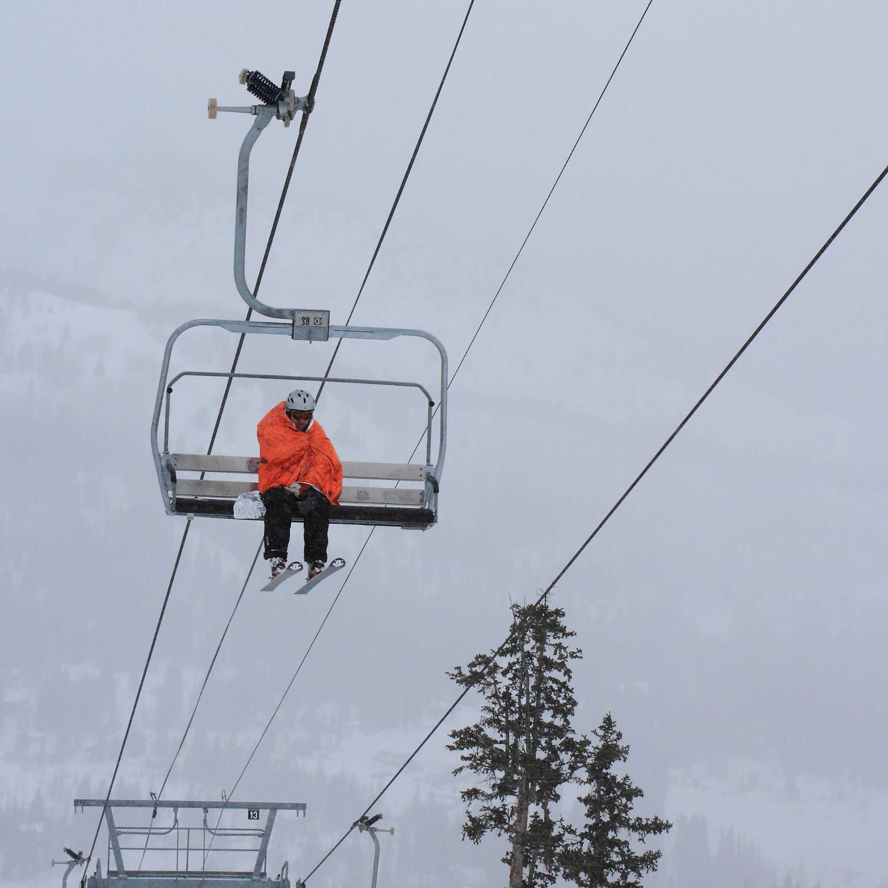 Skier Sitting on Chairlift Wrapped in Orange SOL Emergency Bivvy XL Against a White Sky
