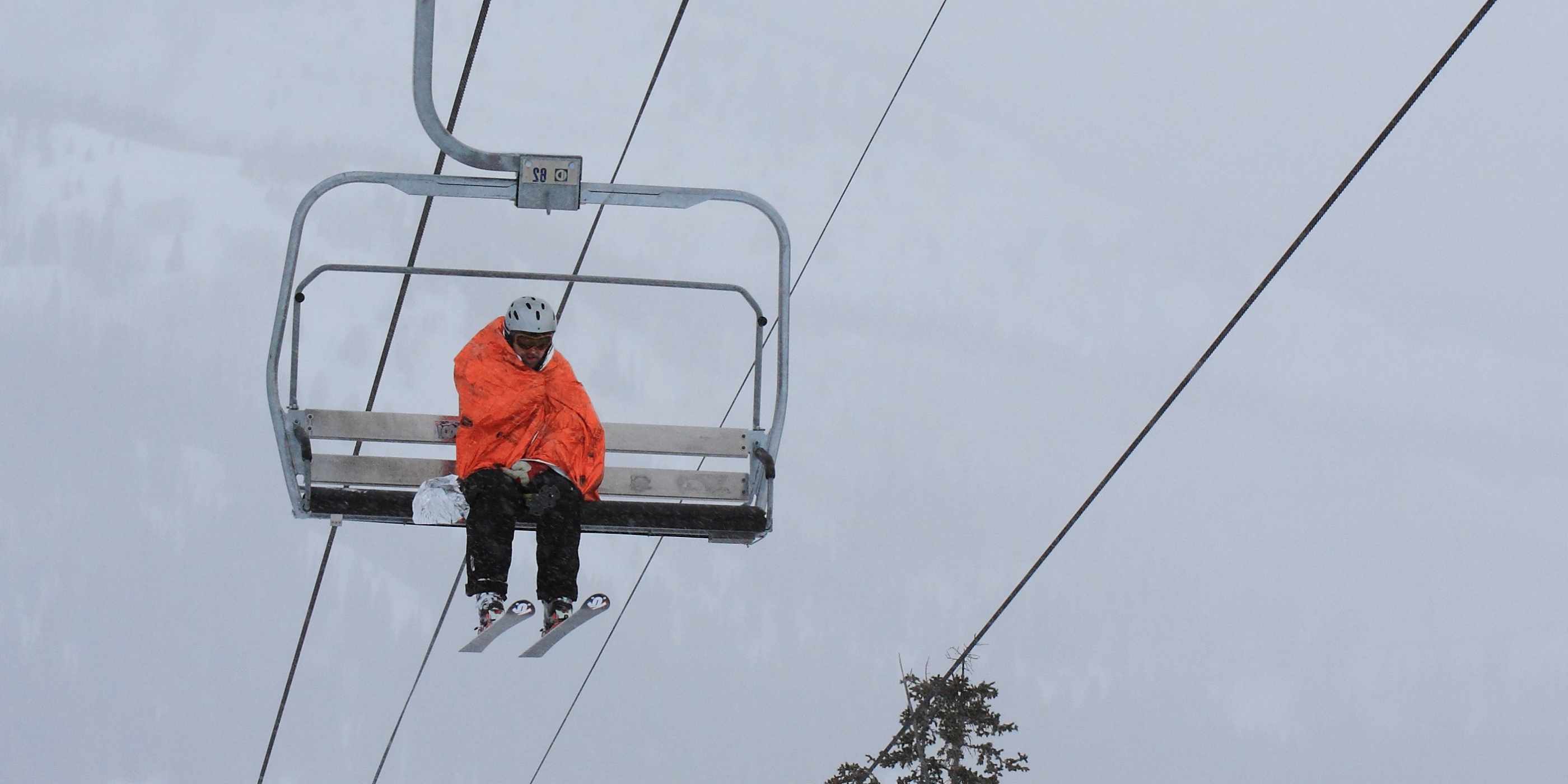 Skier Sitting on Chairlift Wrapped in Orange SOL Emergency Bivvy XL Against a White Sky