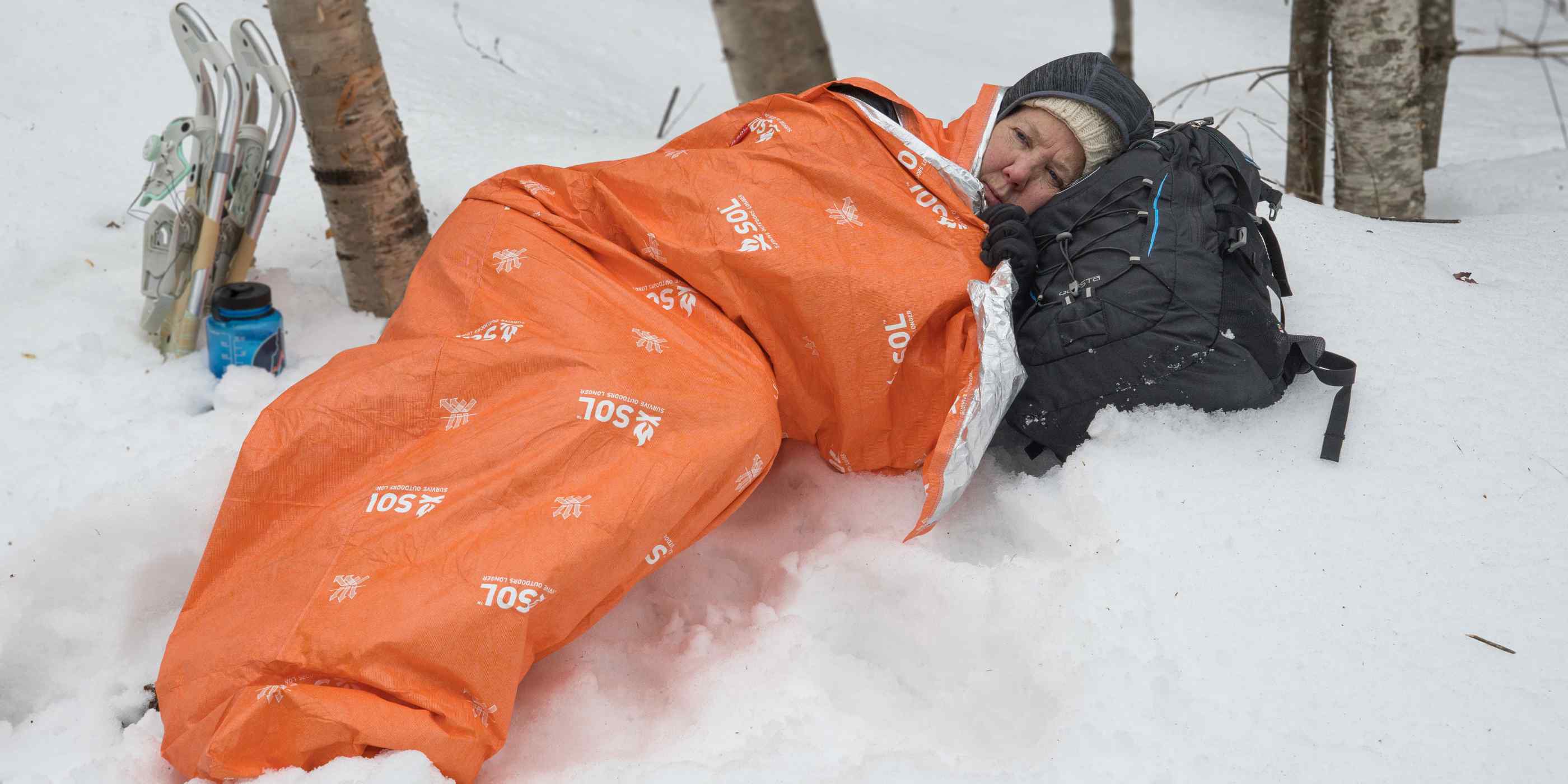 Woman Laying in Snow Wrapped in Orange SOL Escape Lite Bivvy Using Black Backpack as a Pillow and Snowshoes Behind Her