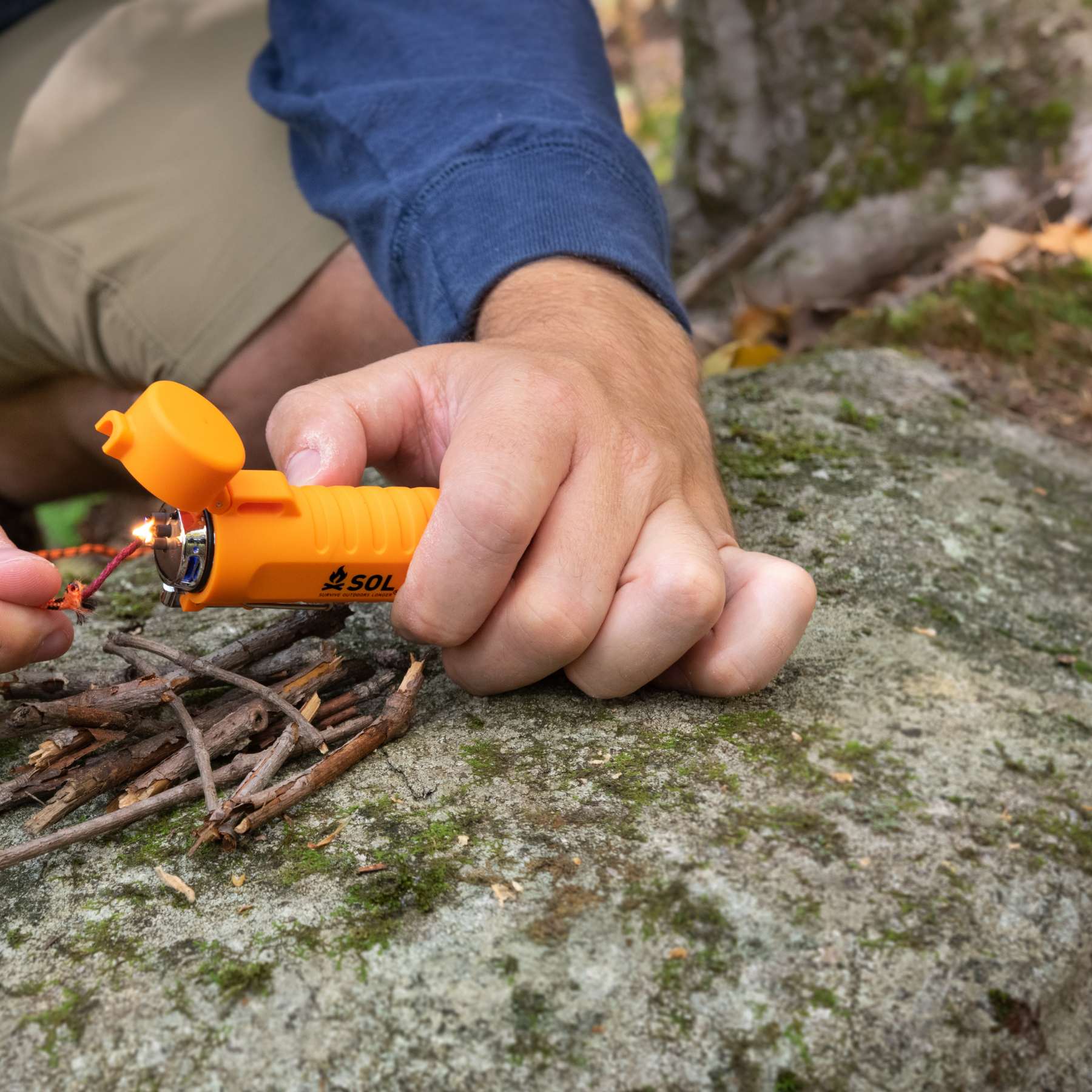 Person Using Orange SOL Fuel Free Lighter Lighting Tinder Cord on Rock with Twigs in a Pile