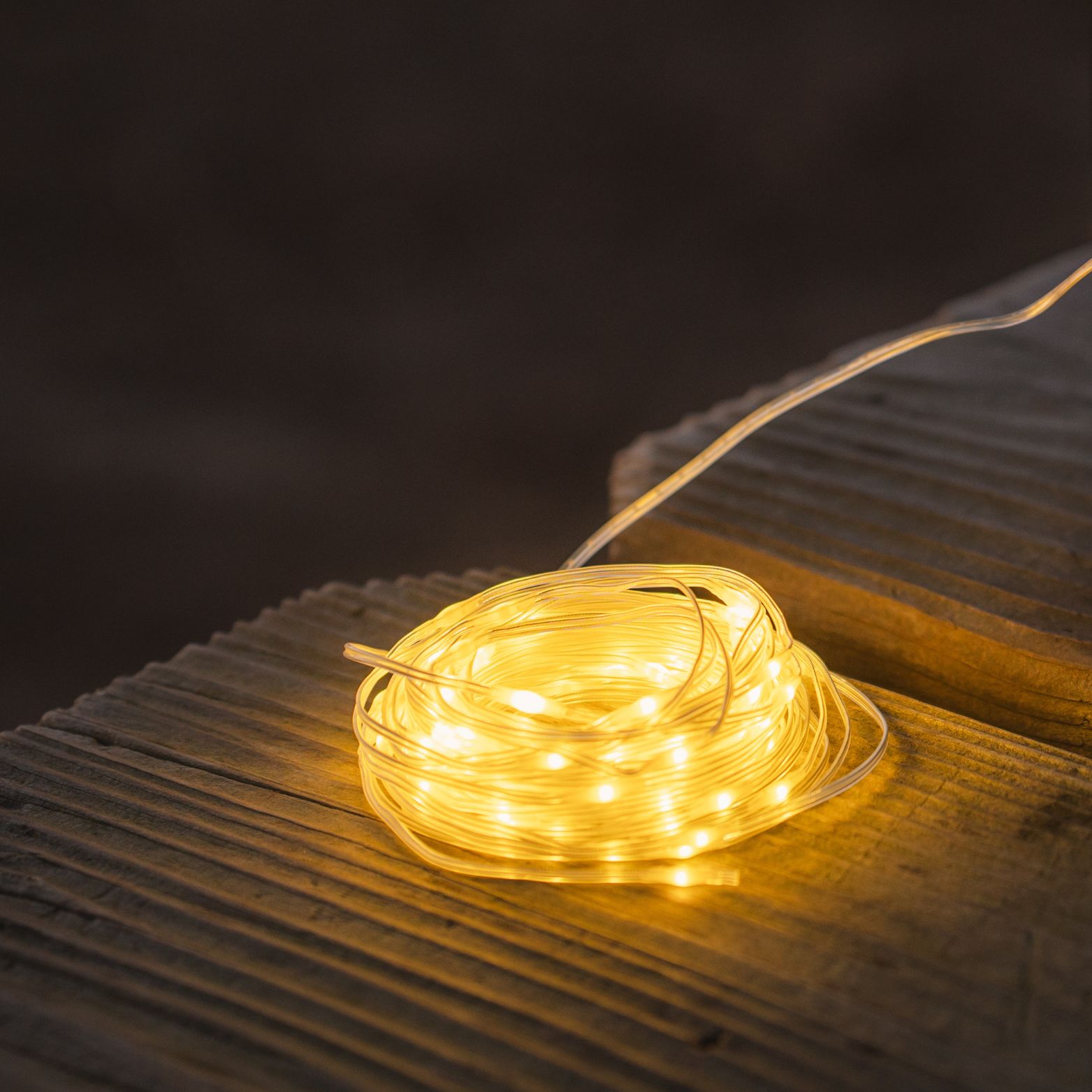A coiled set of LuminAID Solar String Lights glowing warmly on a wooden surface at dusk, casting a soft light against the dark background.