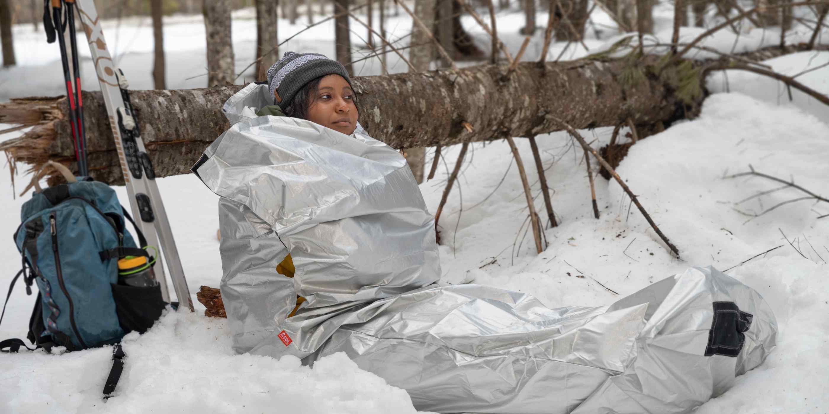 Woman in Silver SOL Thermal Bivvy Sitting in Snow Leaning on Tree with Blue Backpack and Skis Next to Her