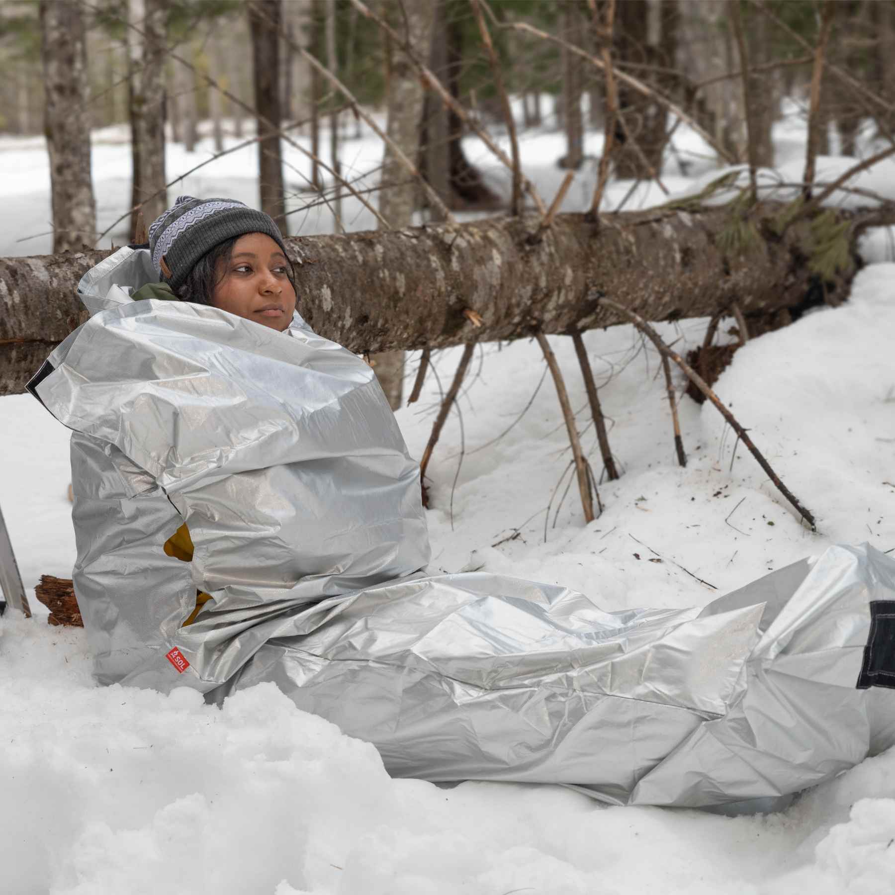 Woman in Silver SOL Thermal Bivvy Sitting in Snow Leaning on Tree with Blue Backpack and Skis Next to Her