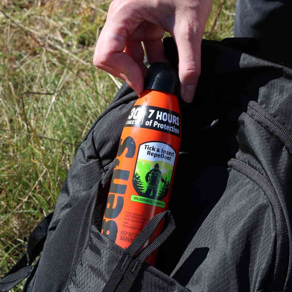 Ben's 30 Tick & Insect Repellent 6 oz. Eco-Spray hand pulling out of black backpack pocket in front of grass