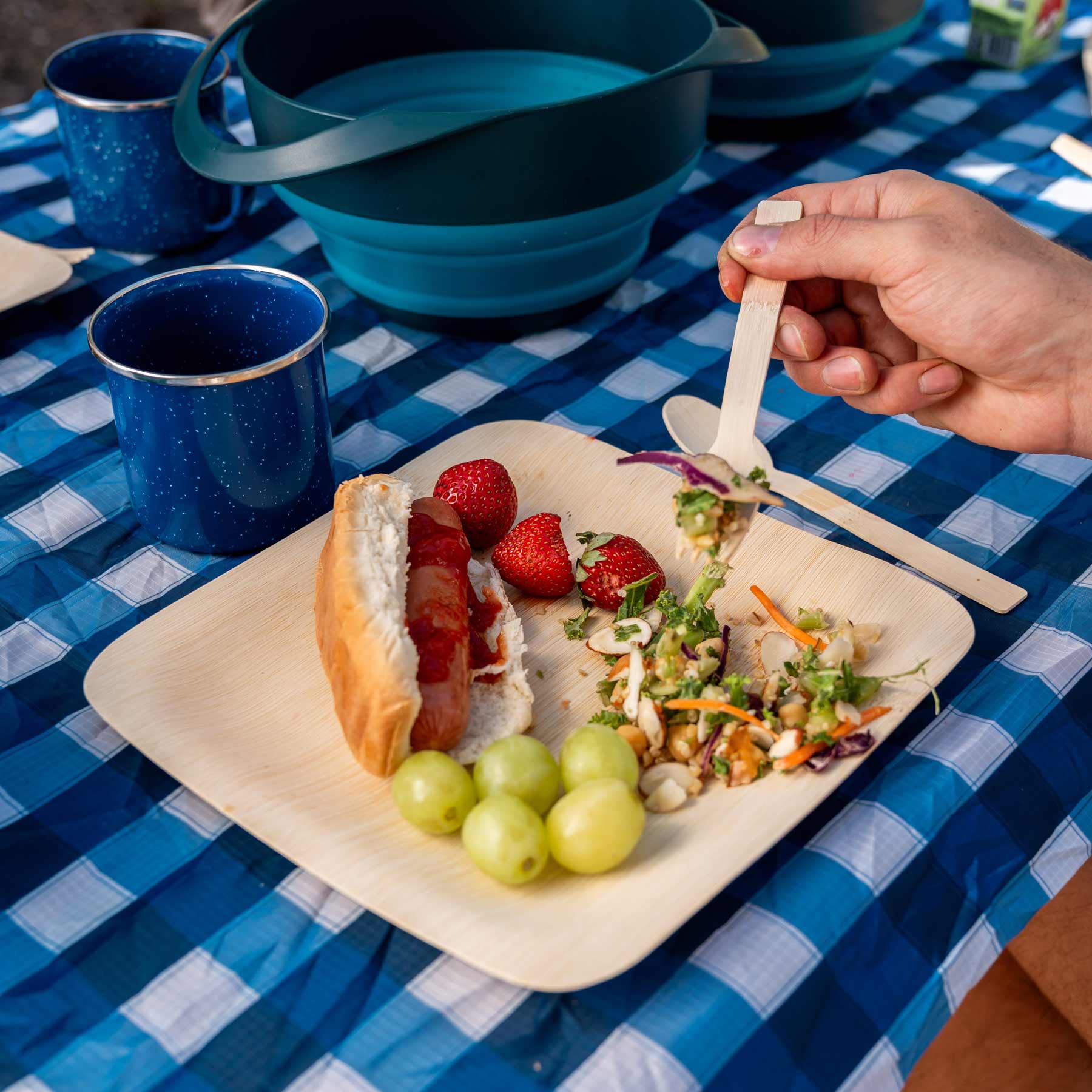 Image shows the Bamboo Dining Set in use on a picnic table. The person using the set is eating salad off the plate using the bamboo fork.
