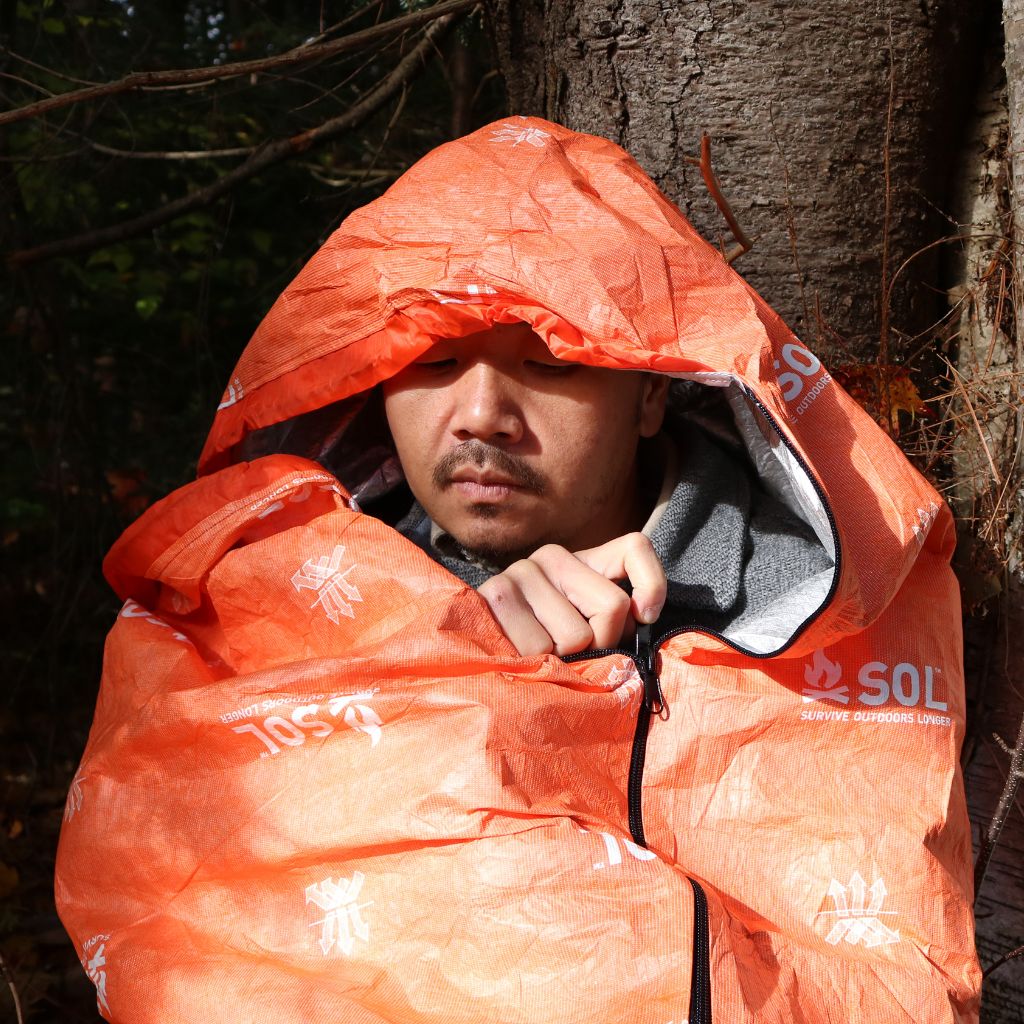 Person wrapped in an orange sleeping bag with SOL branding in a forest setting