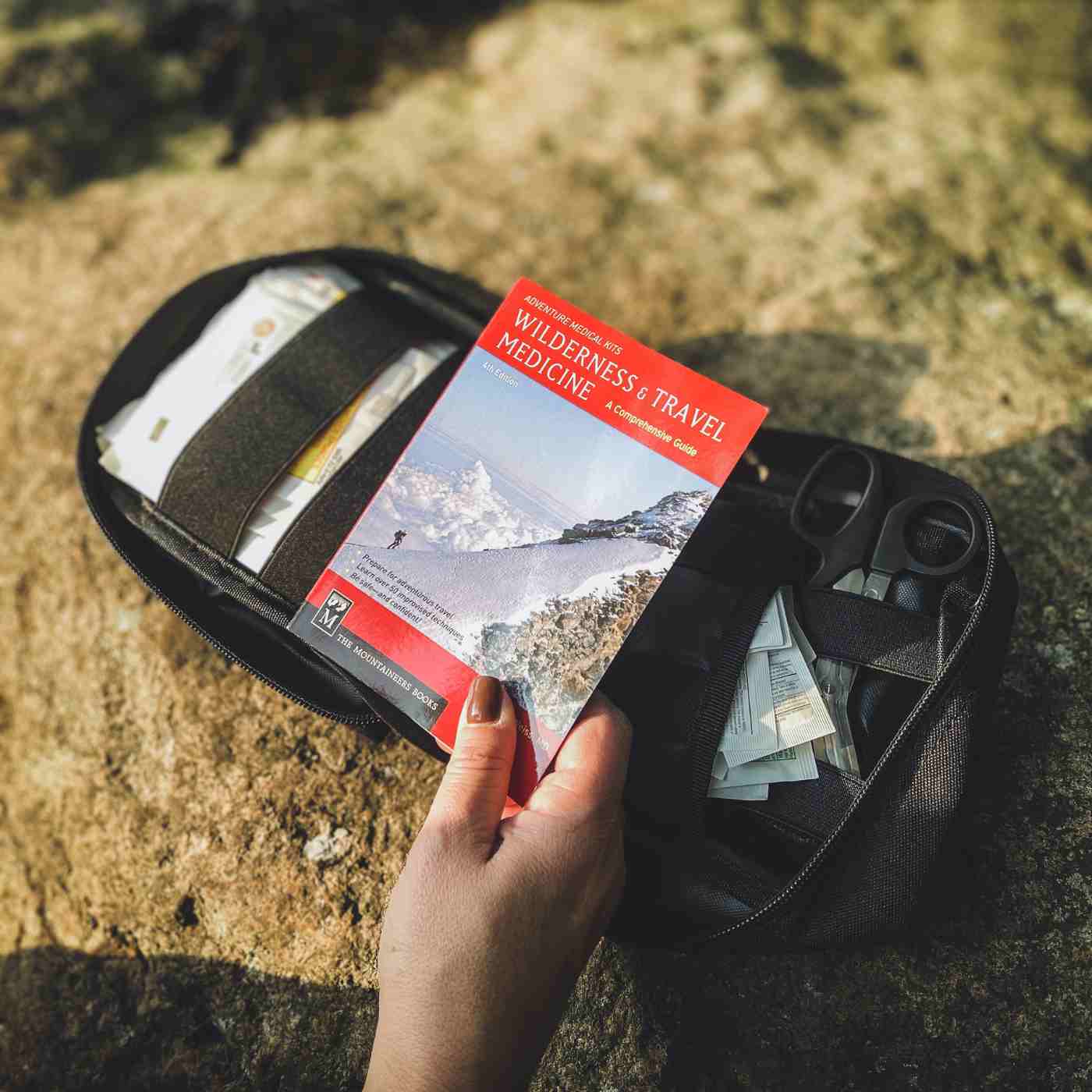 MOLLE Bag Trauma Kit 1.0 - Black person holding Wilderness and Travel Medicine book in front of opened kit on rock