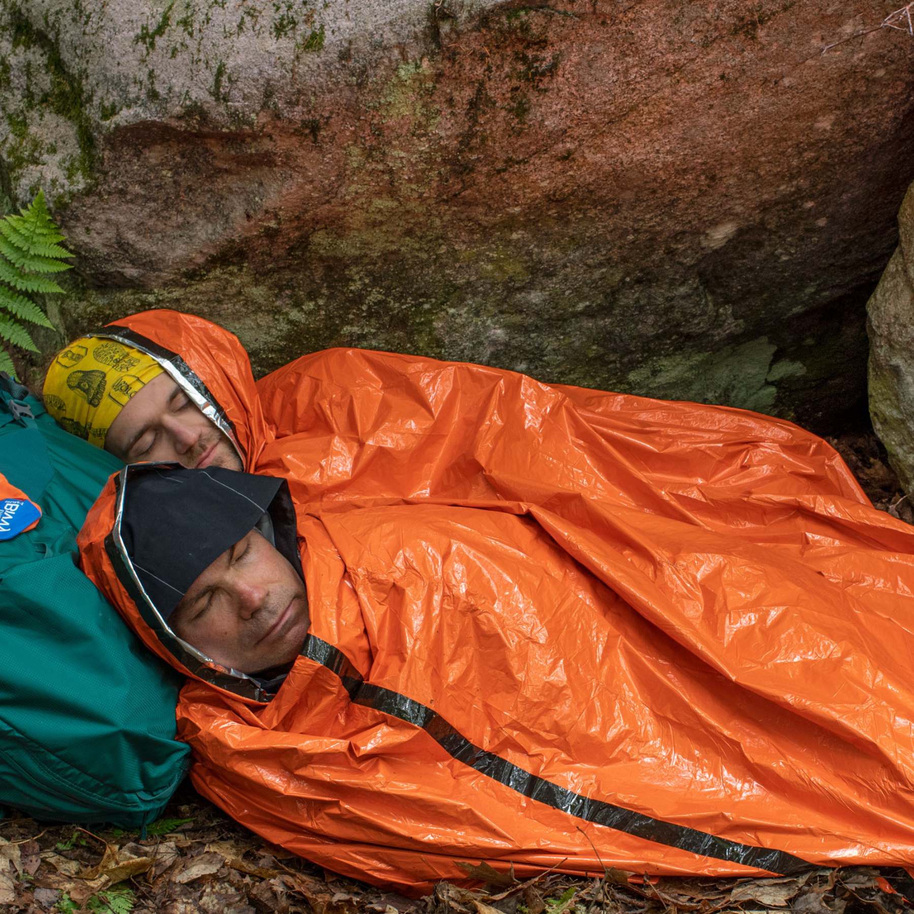 Two Men Wrapped in Orange SOL Emergency XL 2-Person Bivvy on Leaves Using Green Backpack as Pillow