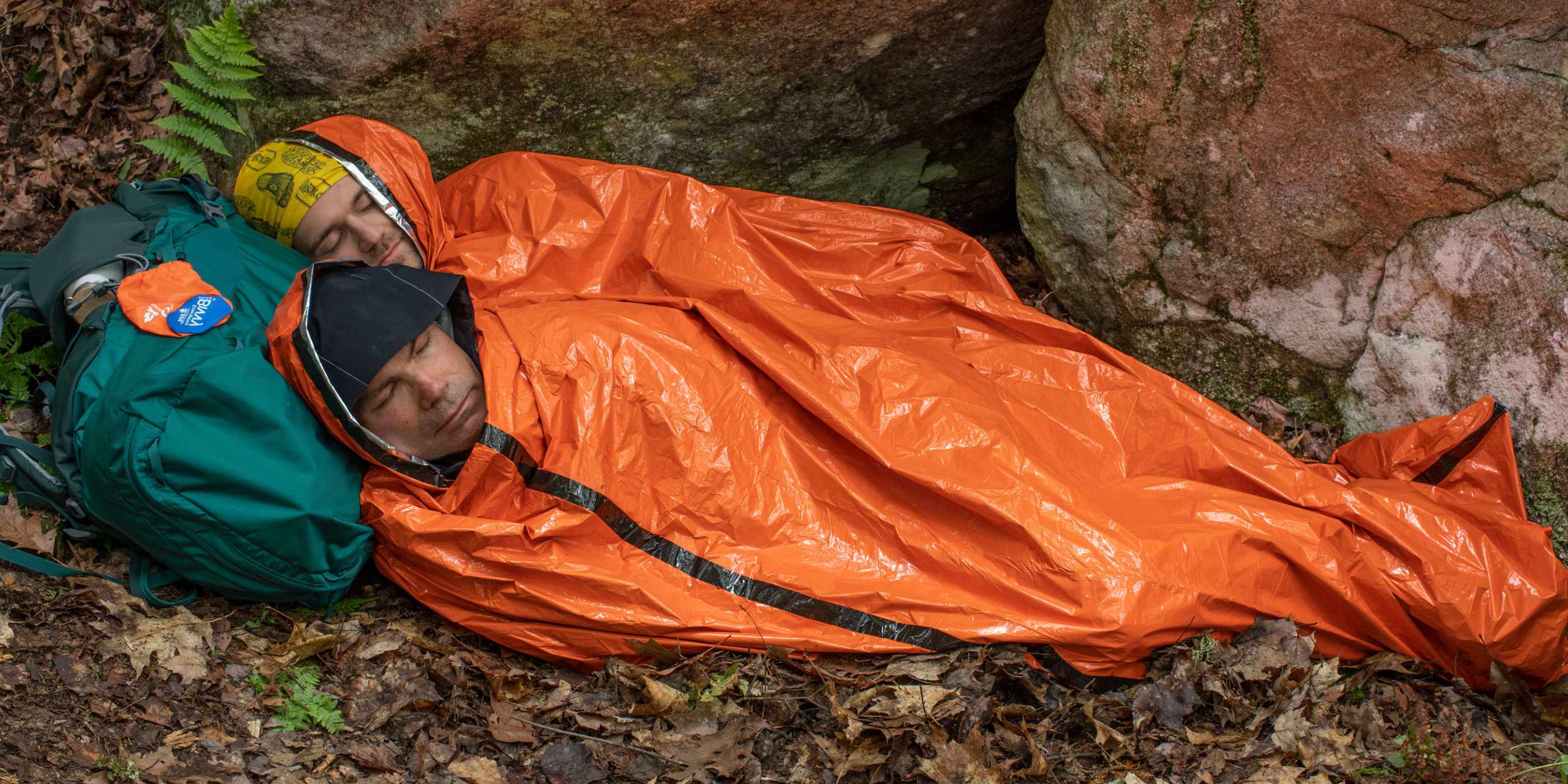 Two Men Wrapped in Orange SOL Emergency XL 2-Person Bivvy on Leaves Using Green Backpack as Pillow