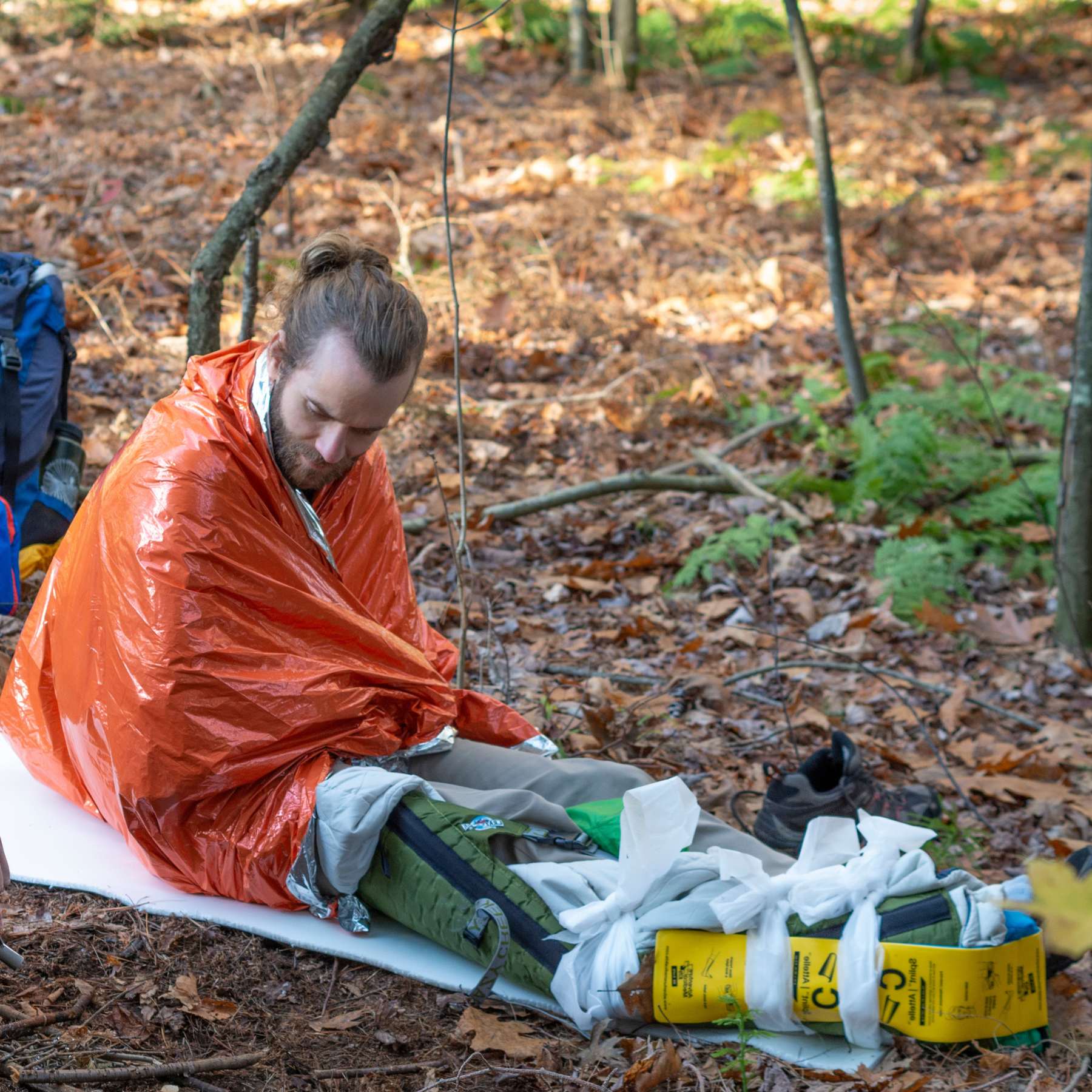 Man Wrapped in Orange SOL Emergency Bivvy Sitting on Leafy Ground