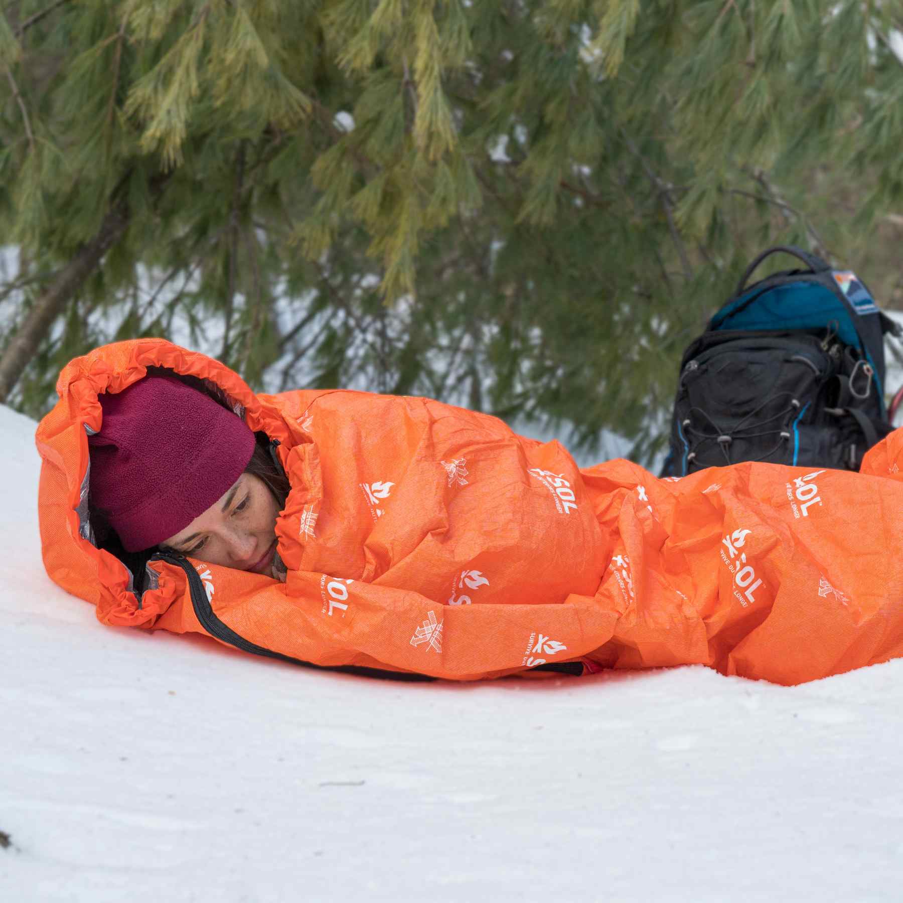 Woman in Red Hat Wrapped in Orange SOL Escape Bivvy Laying on Snow with Backpack and Snowshoes Behind Her