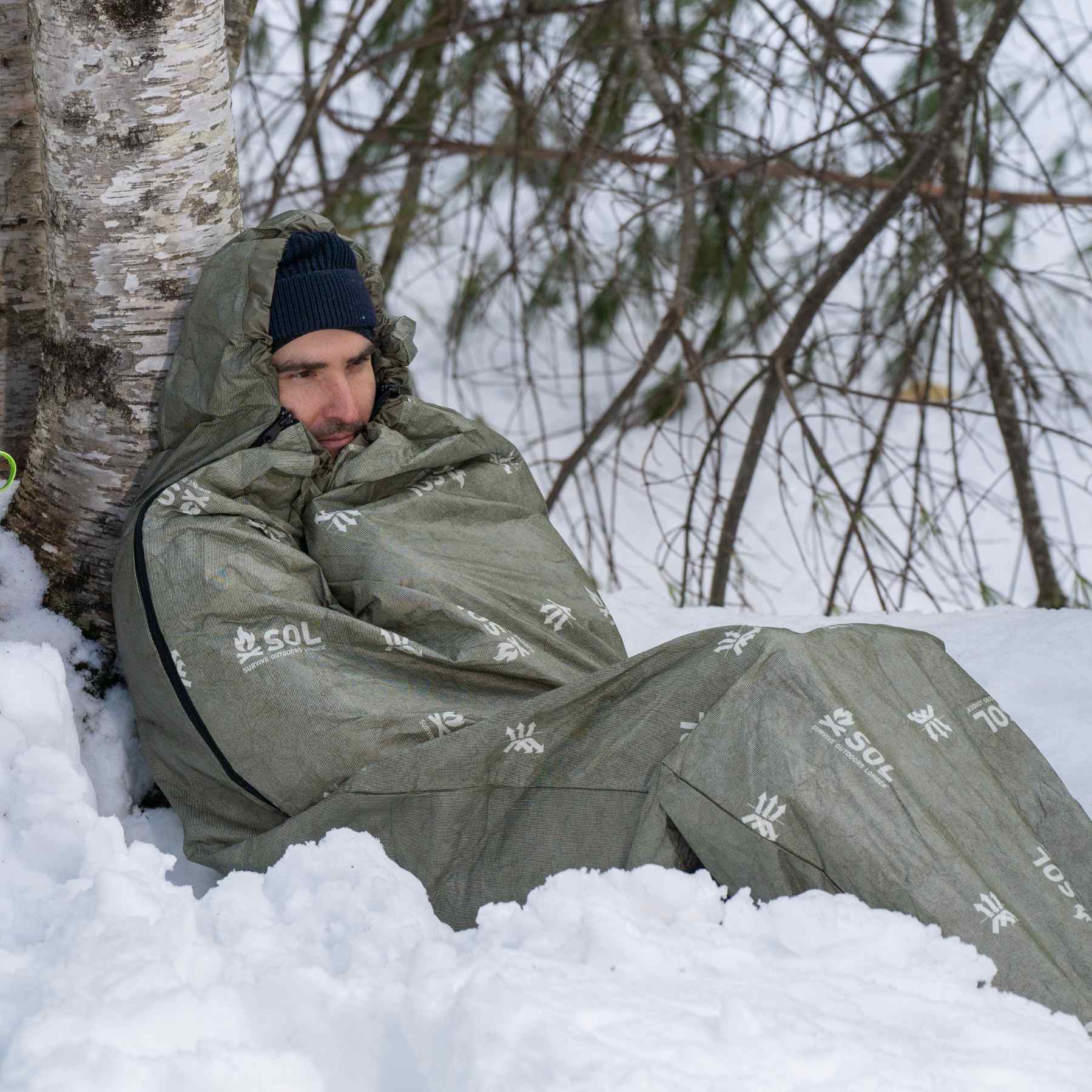 Man Sitting on Snow Leaning on Tree in OD Green SOL Escape Bivvy with Blue Backpack Behind Him