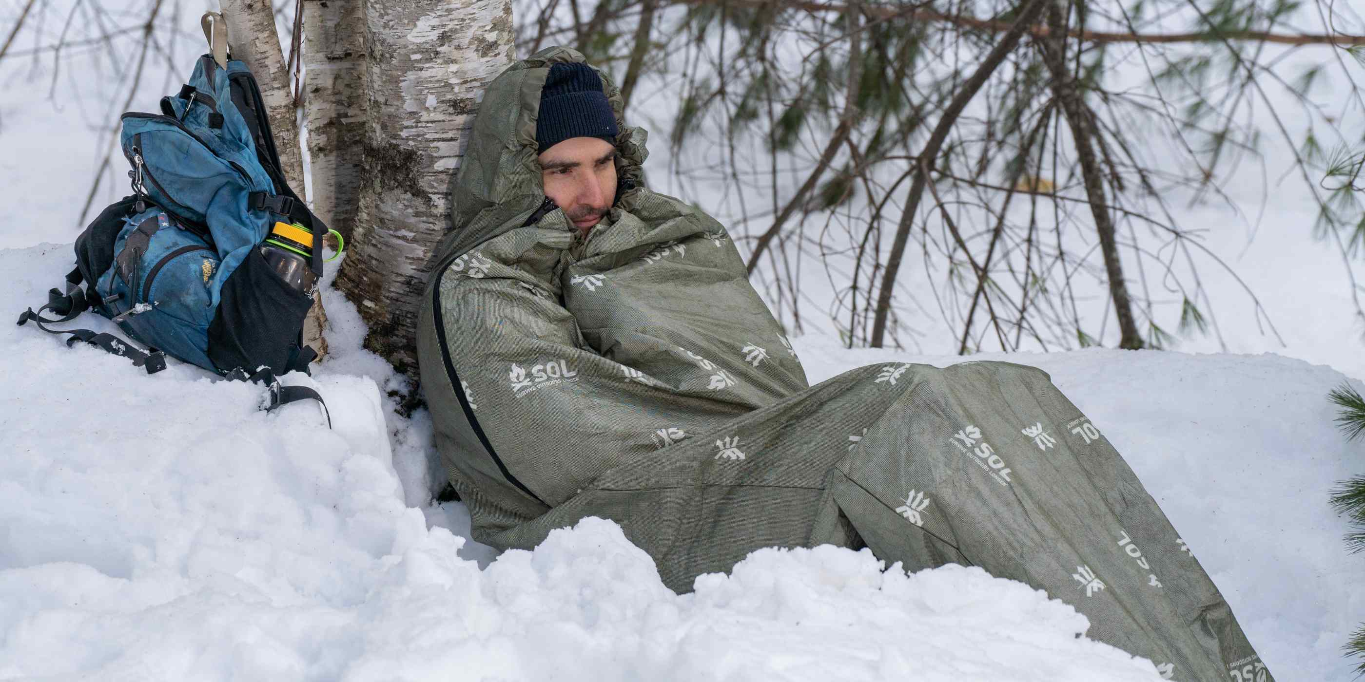 Man Sitting on Snow Leaning on Tree in OD Green SOL Escape Bivvy with Blue Backpack Behind Him