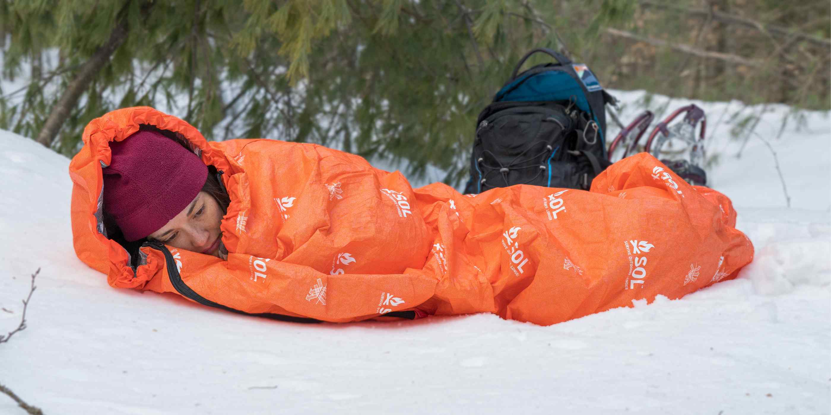 Woman in Red Hat Wrapped in Orange SOL Escape Bivvy Laying on Snow with Backpack and Snowshoes Behind Her