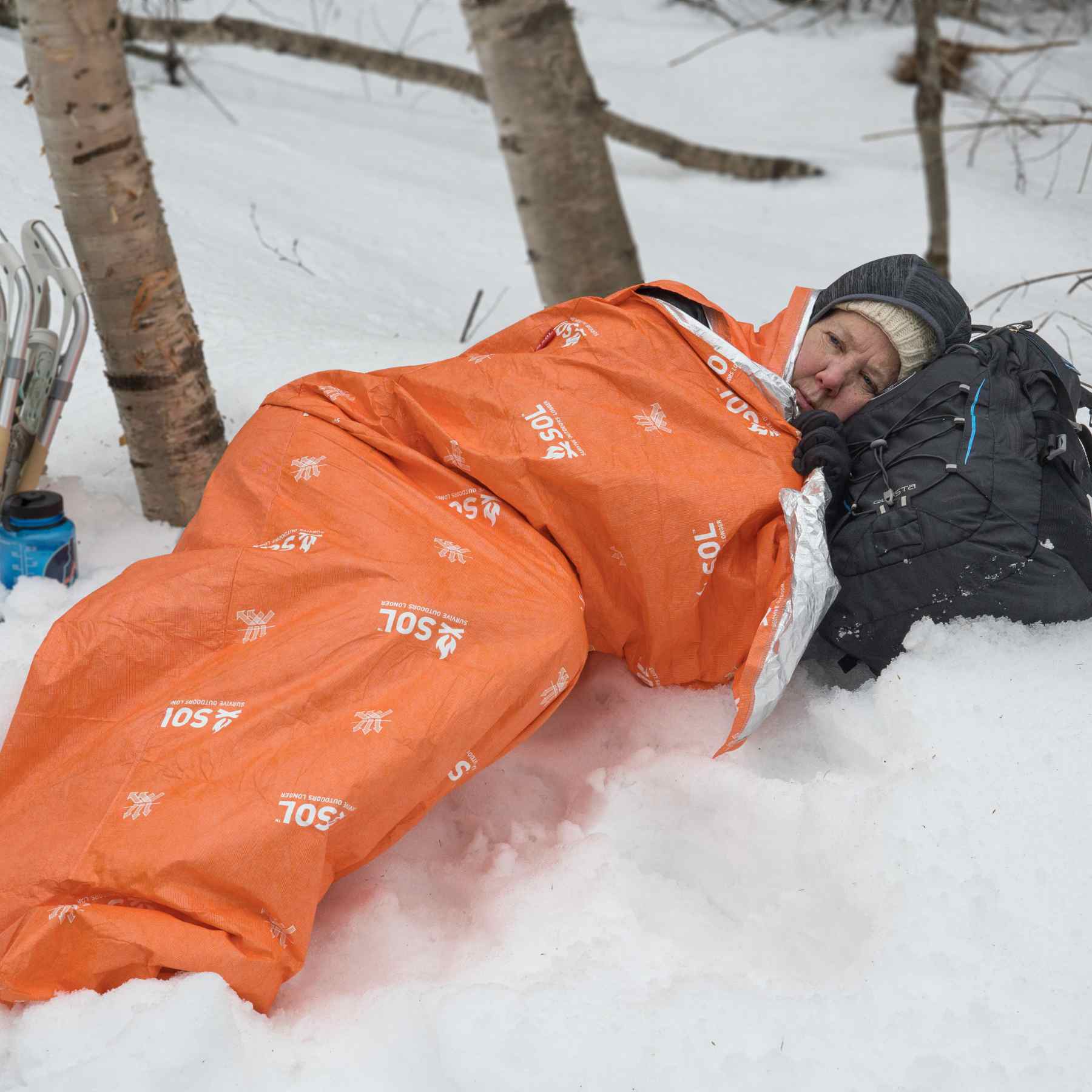 Woman Laying in Snow Wrapped in Orange SOL Escape Lite Bivvy Using Black Backpack as a Pillow and Snowshoes Behind Her