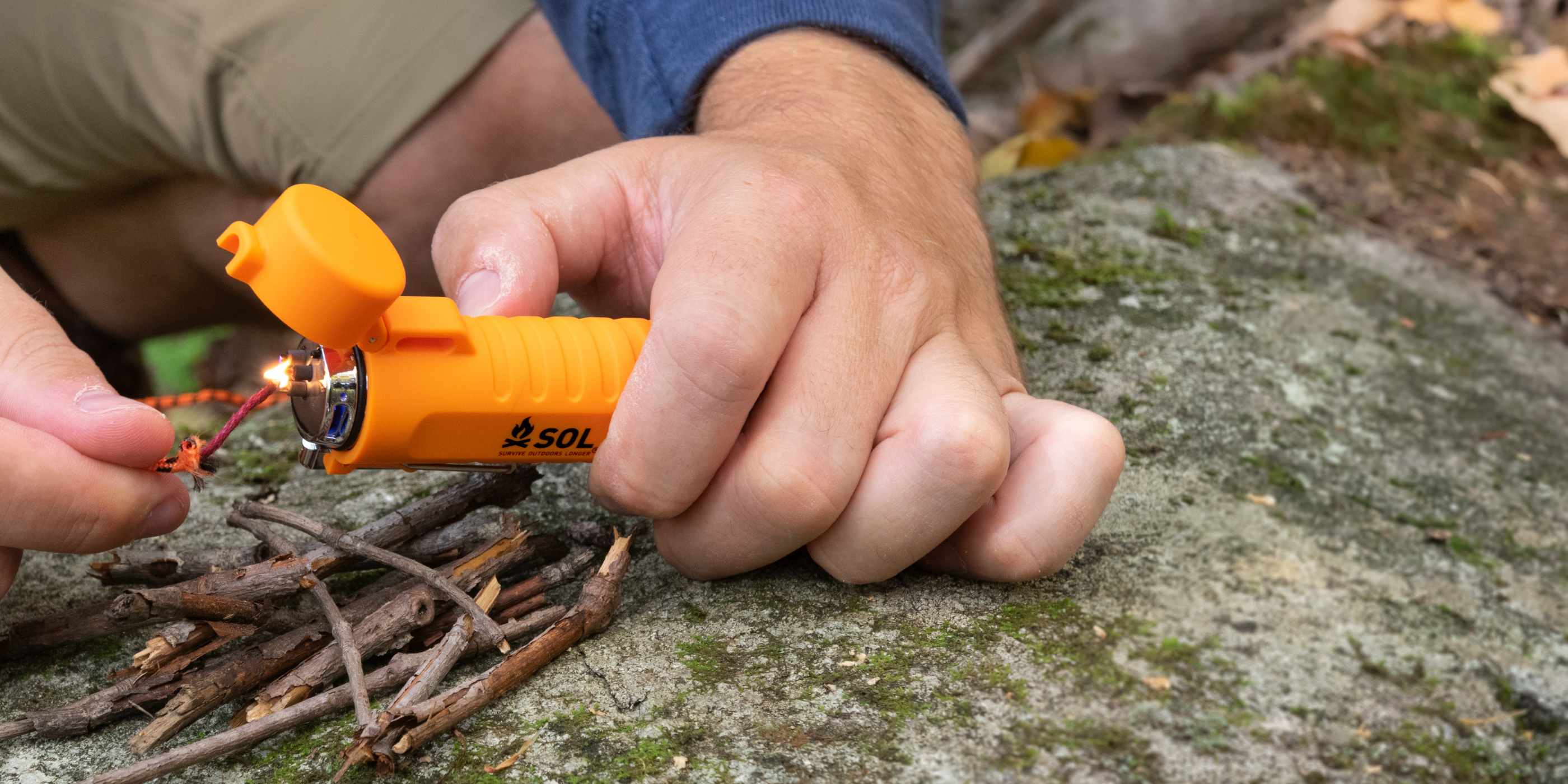 Person Using Orange SOL Fuel Free Lighter Lighting Tinder Cord on Rock with Twigs in a Pile