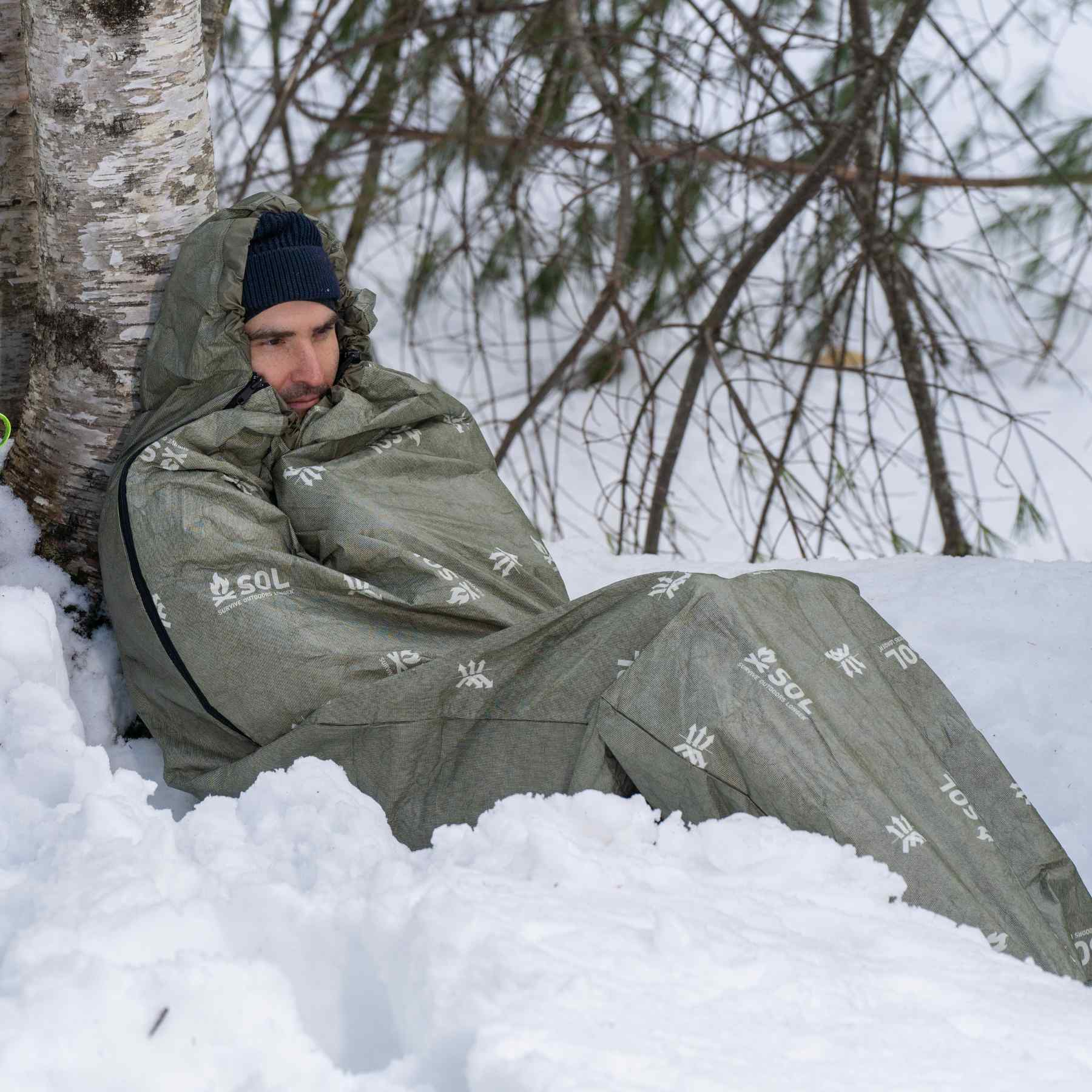 Escape Bivvy OD Green man leaned against tree in snow in bivvy