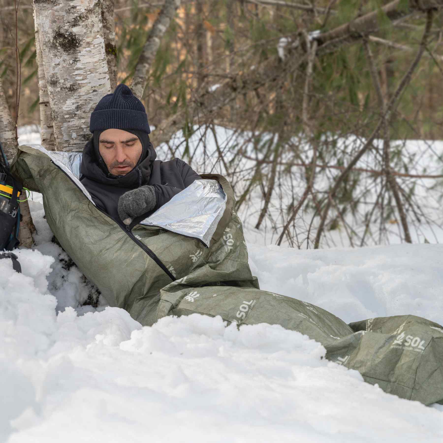 Escape Bivvy OD Green man zipping bivvy in snow while leaned on tree