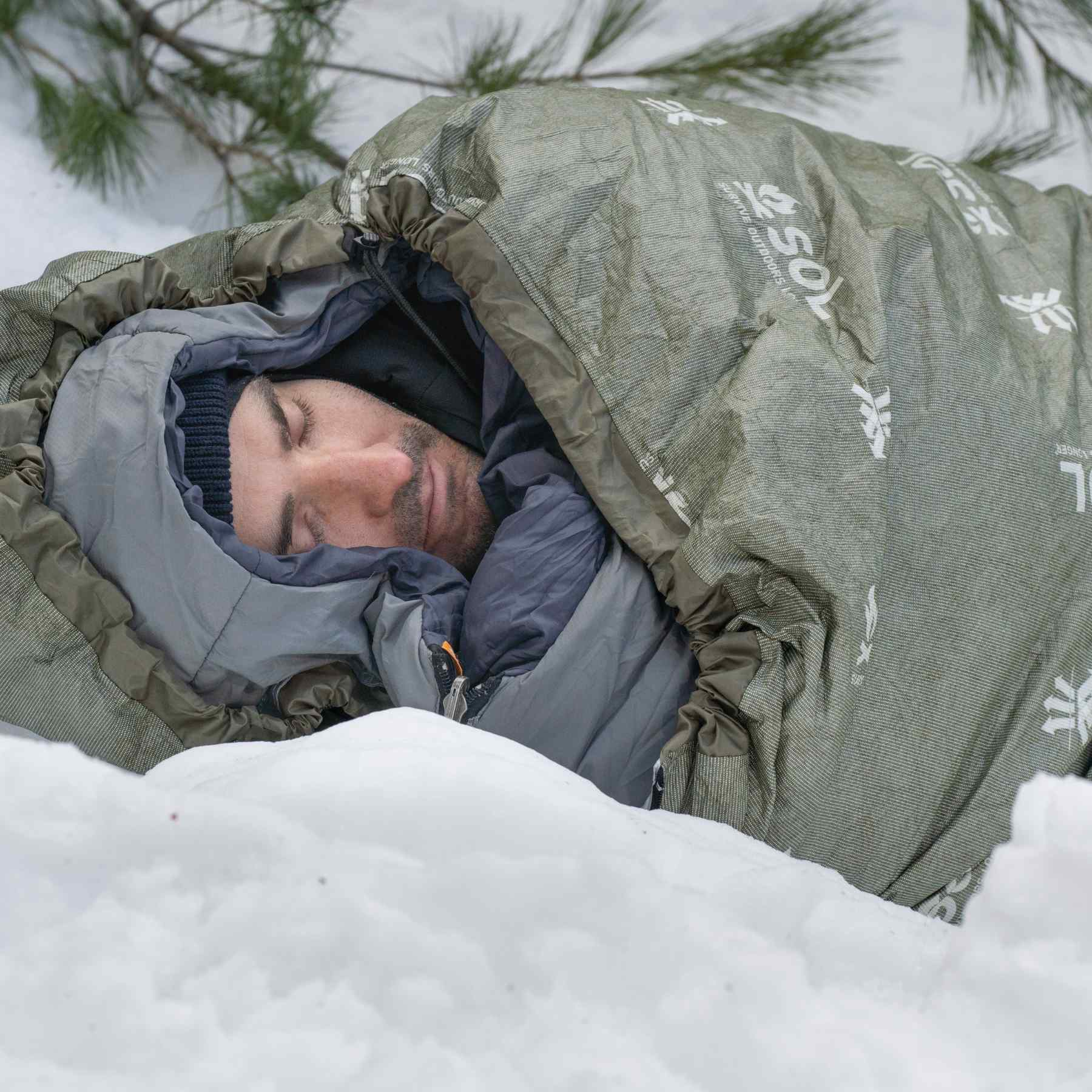 Escape Bivvy OD Green man sleeping on snow in sleeping bag with bivvy on the outside