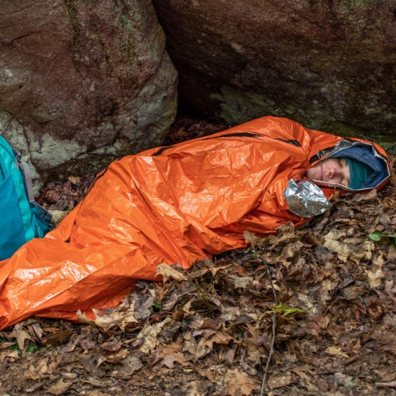 Emergency Bivvy with Rescue Whistle - Orange person sleeping in bivvy on leaves in front of rock