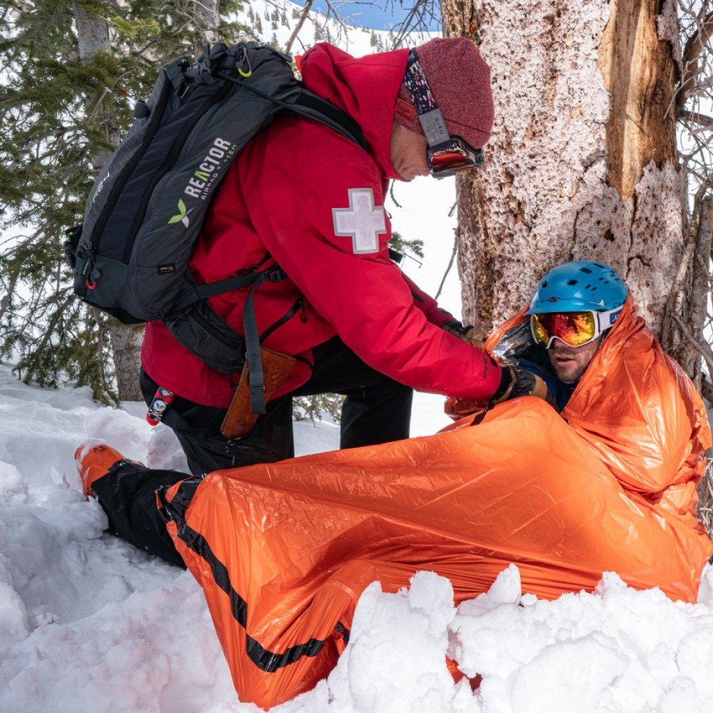 Emergency Bivvy with Rescue Whistle - Orange ski patrol wrapping injured patient in bivvy in snow leaning on tree