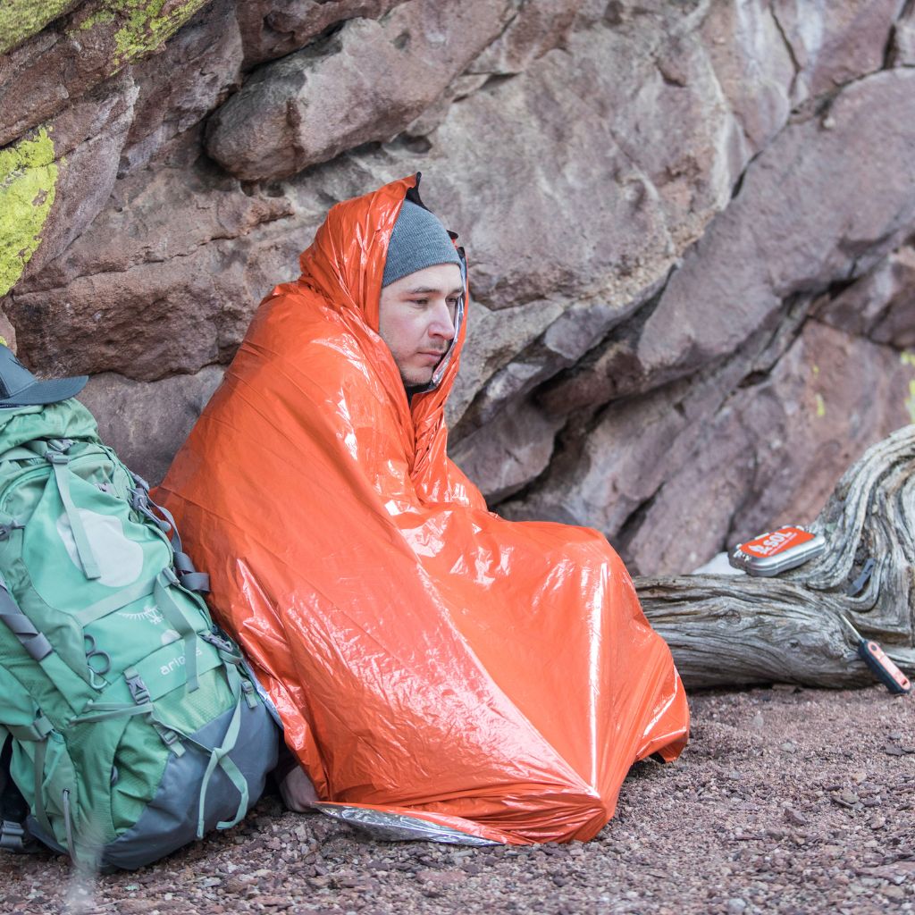 Emergency Blanket man wrapped in blanket in front of a rock next to green backpack
