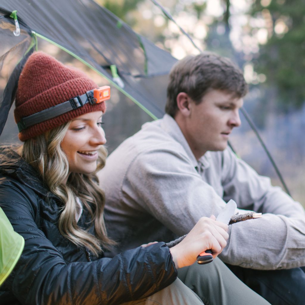 Venture Rechargeable Headlamp on woman's head while sitting in tent next to man while cutting a stick with a knife